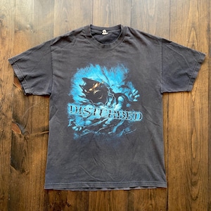 Può includere: Una maglietta nera con una grafica blu e bianca di una creatura con occhi rossi luminosi e la parola "DISTURBED" stampata sotto.