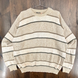 Op de afbeelding: Beige en crèmekleurige trui met horizontale strepen in bruin- en groentinten. De gebreide trui met lange mouwen hangt aan een metalen hanger tegen een houten achtergrond. De trui heeft een ronde hals en geribde manchetten en zoom.