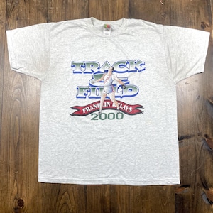 May include: A light grey t-shirt with a graphic promoting a "Track & Field" event. The design includes the text "Track & Field" in blue and green, a runner, and the text "Frank in Relays 2000" in red.