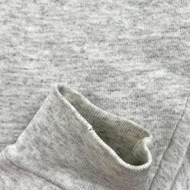 May include: Close-up of a grey sweatshirt sleeve cuff. The cuff is folded over, revealing the inside fabric. The material is a heathered grey, with a ribbed texture on the cuff and a smoother texture on the body of the sleeve.
