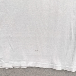 May include: A close-up of a white t-shirt. The shirt has a small stain near the bottom. The fabric appears to be a soft, knit material. The bottom hem is visible.