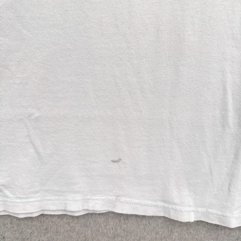 May include: A close-up of a white t-shirt. The shirt has a small stain near the bottom. The fabric appears to be a soft, knit material. The bottom hem is visible.