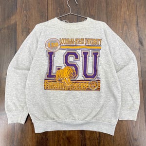 May include: A gray crewneck sweatshirt featuring the Louisiana State University logo. The design includes the letters "LSU" in purple and gold, a tiger graphic, and the words "Fighting Tigers".