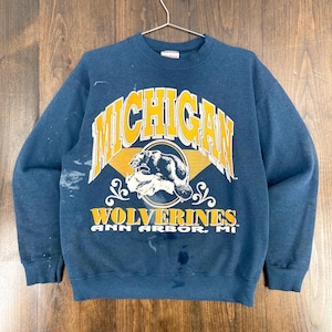 Peut inclure: Un sweat-shirt bleu marine avec le mot "MICHIGAN" en lettres jaunes. En dessous, un dessin de glouton et le mot "WOLVERINES" en jaune. Le texte "ANN ARBOR, MI" est en dessous. Le sweat-shirt est suspendu à un cintre.