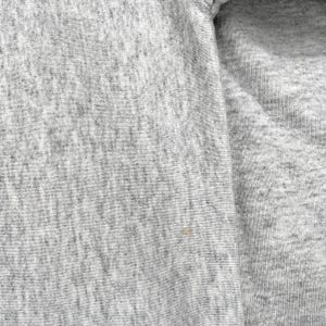 May include: Close-up of a heather grey fabric, possibly a sweatshirt. The texture is evident, with subtle variations in the grey shades. The image shows a section of the garment, including a seam.