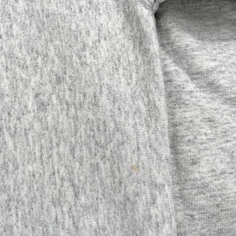 May include: Close-up of a heather grey fabric, possibly a sweatshirt. The texture is evident, with subtle variations in the grey shades. The image shows a section of the garment, including a seam.