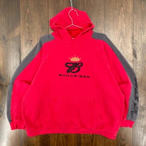 May include: A red hoodie with grey sleeves. The front displays the Budweiser logo, a crown, and the word "BUDWEISER" in black. The right sleeve has the text "DALE JR" in grey.