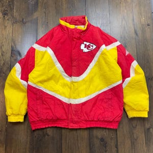 May include: A vintage Kansas City Chiefs jacket in red, white and yellow with a team logo on the front. The jacket has a zip fastening and a stand-up collar.