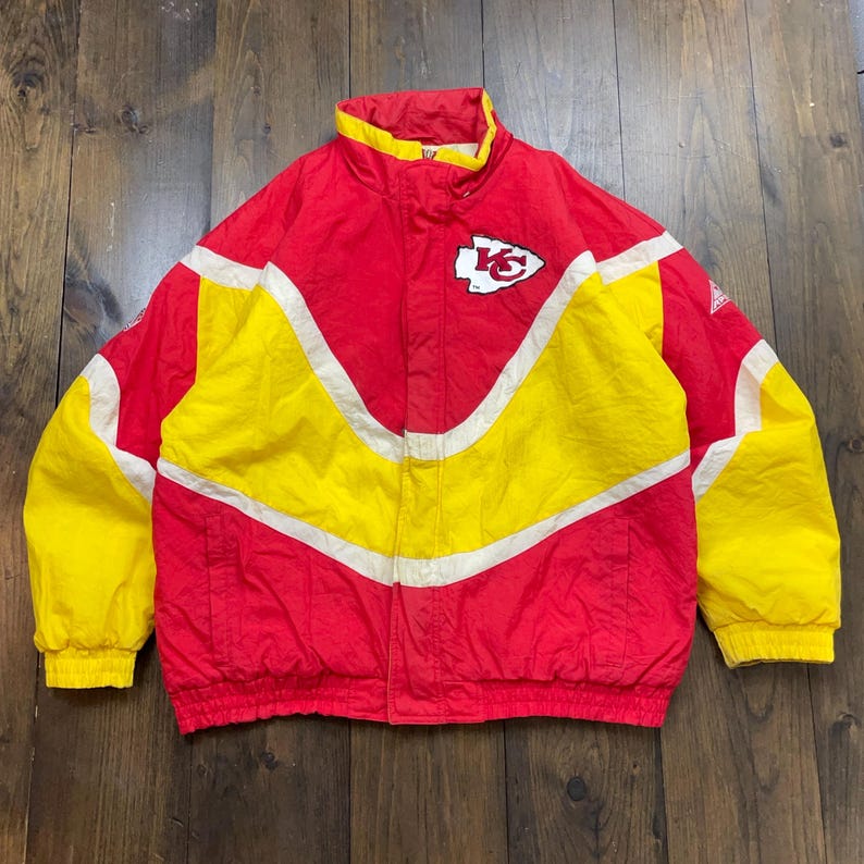May include: A vintage Kansas City Chiefs jacket in red, white and yellow with a team logo on the front. The jacket has a zip fastening and a stand-up collar.