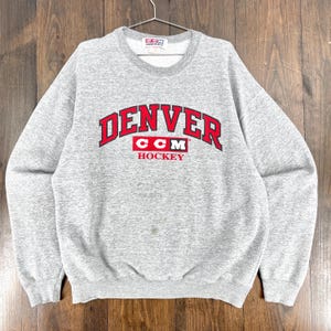 Puede incluir: Una sudadera gris de cuello redondo con la palabra "DENVER" en letras rojas arqueadas. Debajo, el texto "CCM HOCKEY" también en rojo. La sudadera está colgada de una percha metálica.
