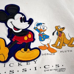 May include: White sweatshirt featuring Mickey Mouse, Donald Duck, and Pluto appliqu&eacute;s. Mickey is in red shorts and yellow shoes. Donald Duck is in a blue sailor suit. Pluto is a yellow dog. The text "MICKEY S.S.I.C.S." is at the bottom.