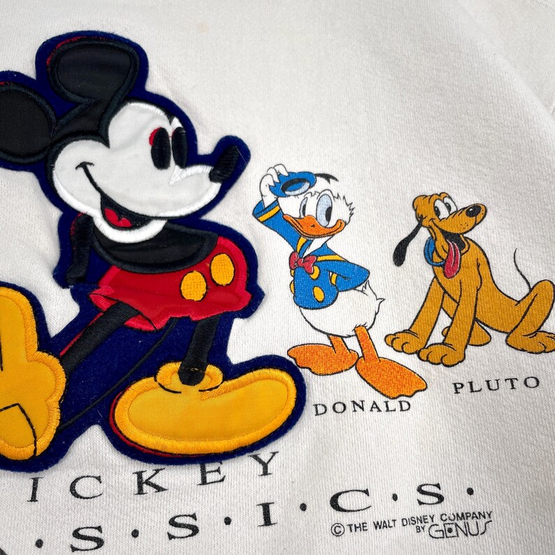 May include: White sweatshirt featuring Mickey Mouse, Donald Duck, and Pluto appliqu&eacute;s. Mickey is in red shorts and yellow shoes. Donald Duck is in a blue sailor suit. Pluto is a yellow dog. The text "MICKEY S.S.I.C.S." is at the bottom.
