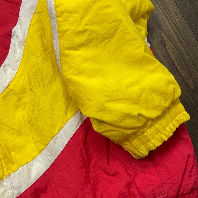 May include: A red, white and yellow windbreaker jacket with a zip closure. The jacket has a ribbed cuff and a white stripe down the sleeve.