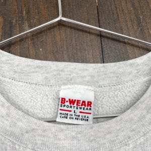 May include: Close-up of a grey sweatshirt with a label reading "B-WEAR SPORTSWEAR" in red and white. The label also states "MADE IN THE U.S.A." and "CARE ON REVERSE". The sweatshirt is hanging on a metal hanger.