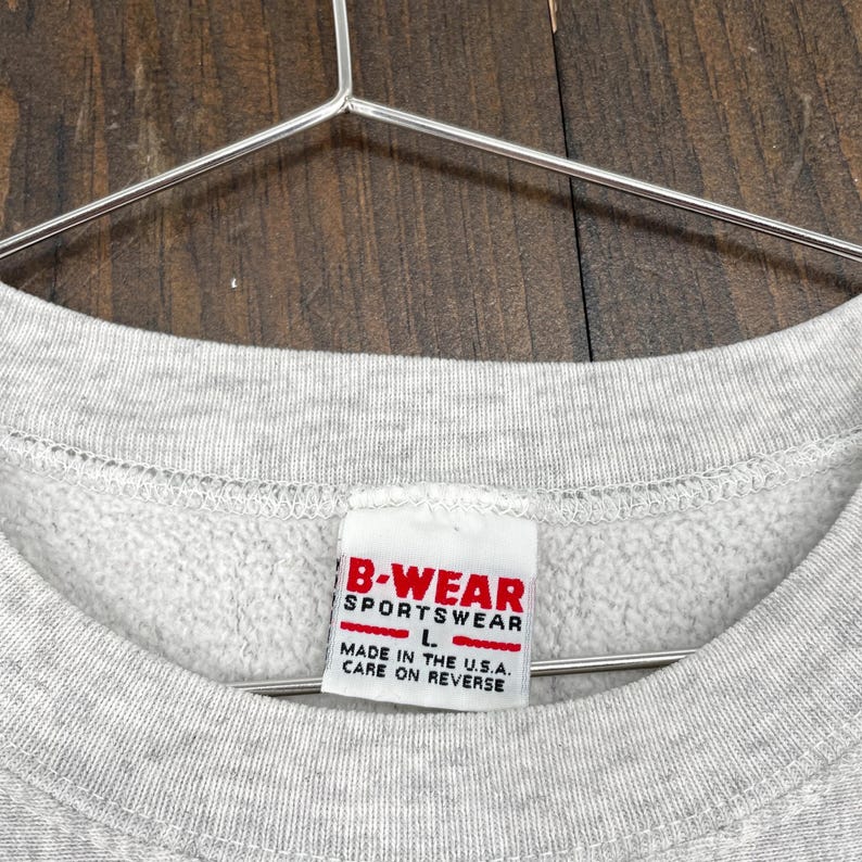 May include: Close-up of a grey sweatshirt with a label reading "B-WEAR SPORTSWEAR" in red and white. The label also states "MADE IN THE U.S.A." and "CARE ON REVERSE". The sweatshirt is hanging on a metal hanger.