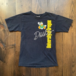 Puede incluir: Camiseta negra con un gráfico de pato de dibujos animados amarillo y verde y el texto "University of Oregon" y "Duck" en amarillo.