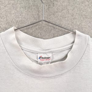 May include: Close-up of a light gray t-shirt with a crew neck collar. The shirt is hanging on a metal hanger. A white tag with the brand name "Stedman" is visible near the collar.
