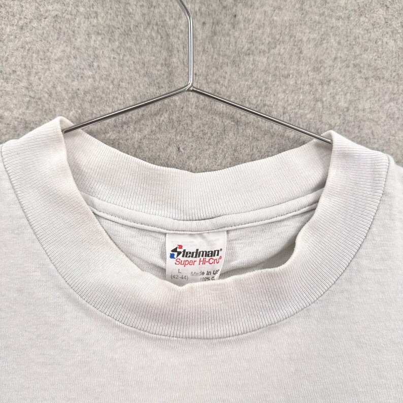May include: Close-up of a light gray t-shirt with a crew neck collar. The shirt is hanging on a metal hanger. A white tag with the brand name "Stedman" is visible near the collar.