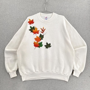 May include: A white crewneck sweatshirt featuring a graphic print of autumn maple leaves in shades of orange, red, and dark green. The leaves are arranged in a cluster on the upper left side of the chest, with a single dark green leaf on the right.