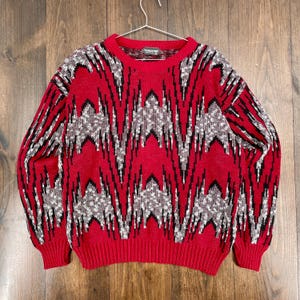 May include: A red sweater with a geometric pattern in black and white. The sweater has a crew neck and ribbed cuffs and hem. The pattern is a zig-zag design. The sweater is hanging on a metal hanger.