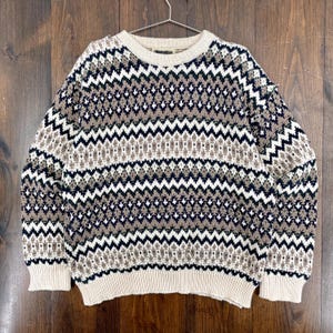 Op de afbeelding: Een beige, marineblauwe, witte en bruine trui met een patroon en een ronde hals. De trui heeft een zich herhalend zigzagontwerp. De manchetten en zoom zijn effen beige. De trui hangt aan een metalen hanger.