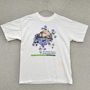 May include: White t-shirt featuring a graphic of a tabby cat resting among purple flowers. The shirt includes the text "The Humane Society of the United States" and "MEMBER 2001" in green and black.