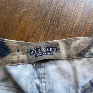 May include: A close-up of a brown and tan camouflage fabric with a brown label that reads "JERZEES Outdoors" in white text.