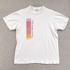 May include: White t-shirt with the IBM logo and a vertical bar code design in red, orange, and yellow. Text on the shirt reads "I Passed the IBM Bar Exam!" and "9 of 9". The shirt is hanging on a metal hanger.