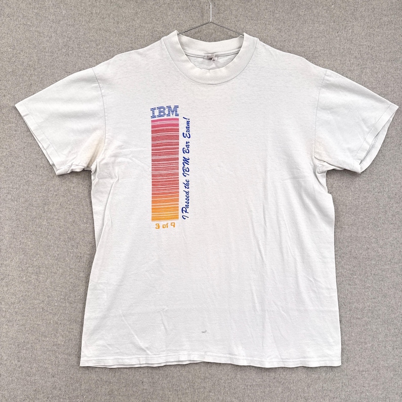 May include: White t-shirt with the IBM logo and a vertical bar code design in red, orange, and yellow. Text on the shirt reads "I Passed the IBM Bar Exam!" and "9 of 9". The shirt is hanging on a metal hanger.