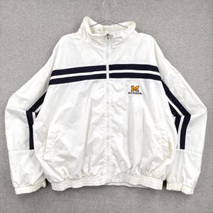 May include: White zip-up jacket with navy blue stripes across the chest and sleeves. Features the word "MICHIGAN" in gold lettering. The jacket has a high collar and a ribbed hem and cuffs.