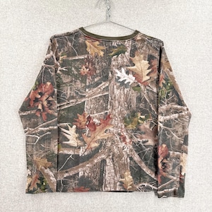 Pode incluir: Uma camisa de manga comprida com um padr&atilde;o de camuflagem florestal realista em tons de castanho, verde e bege. O design apresenta texturas detalhadas de casca de &aacute;rvore, ramos e folhas de carvalho ca&iacute;das por todo o tecido, com uma gola redonda em verde azeitona.