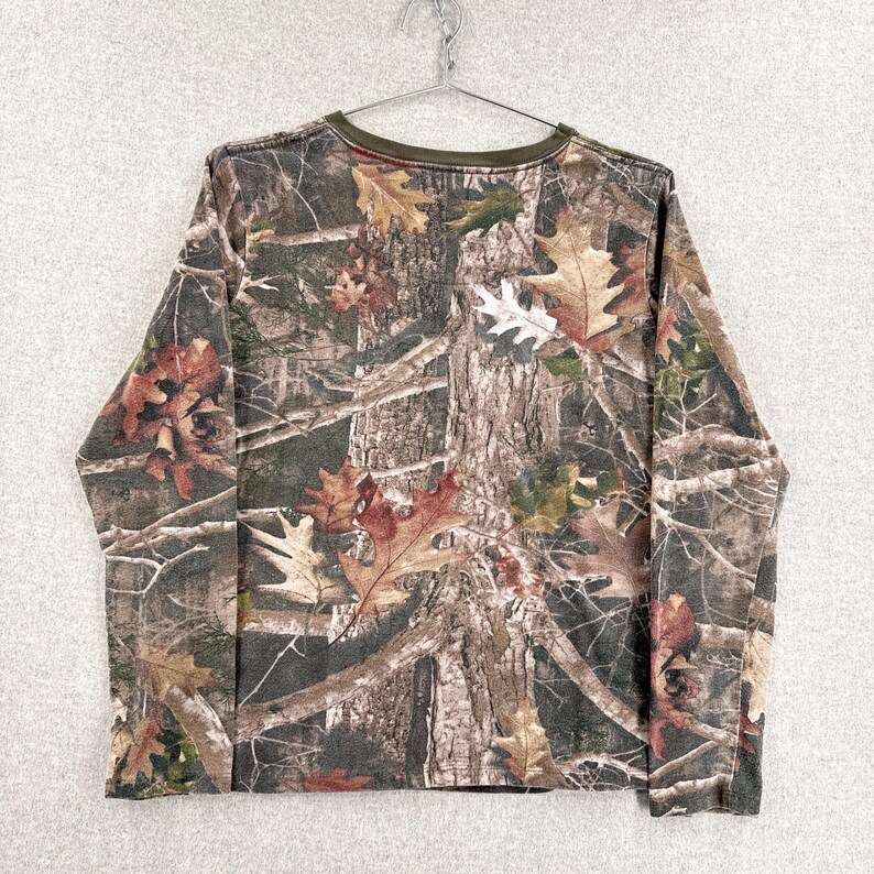 Pode incluir: Uma camisa de manga comprida com um padr&atilde;o de camuflagem florestal realista em tons de castanho, verde e bege. O design apresenta texturas detalhadas de casca de &aacute;rvore, ramos e folhas de carvalho ca&iacute;das por todo o tecido, com uma gola redonda em verde azeitona.
