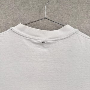 May include: Close-up of a light gray t-shirt hanging on a metal hanger. The shirt has a crew neck and shows signs of wear, including a small hole near the collar and several small dark spots on the fabric.