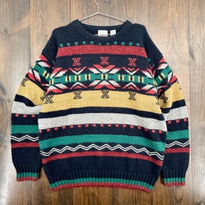May include: A navy blue sweater with a crew neck and long sleeves. The sweater features a colorful geometric pattern in red, green, yellow, and white. The design includes horizontal stripes and zig-zag patterns. The sweater is hanging on a metal hanger.