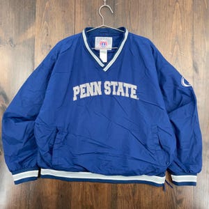 Puede incluir: Un cortavientos azul de Penn State con cuello en V y ribete blanco y gris. Las palabras "PENN STATE" están en letras grises en el pecho. La chaqueta tiene un cordón en la parte inferior y un pequeño logotipo en la manga.