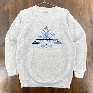 May include: A light grey crew neck sweatshirt. The front displays a blue and white graphic with the words "NORTH CAROLINA" and "TAR HEELS". The sweatshirt is suspended from a metal hanger.