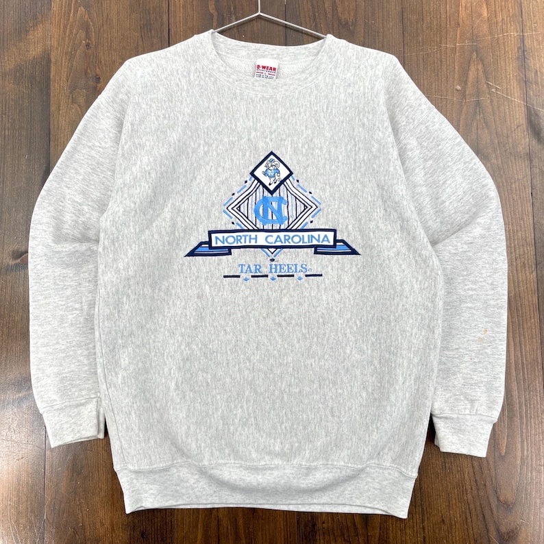 May include: A light grey crew neck sweatshirt. The front displays a blue and white graphic with the words "NORTH CAROLINA" and "TAR HEELS". The sweatshirt is suspended from a metal hanger.