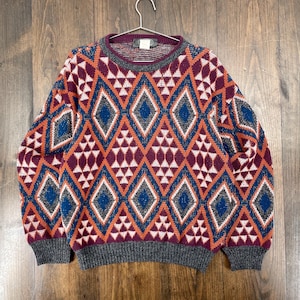 May include: A patterned sweater with a geometric design. The sweater features a repeating pattern of diamonds and triangles in shades of orange, blue, white, and burgundy. The collar, cuffs, and hem are gray. The sweater is hanging on a metal hanger.