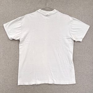 May include: A plain white short-sleeved t-shirt hanging on a metal hanger. The shirt is made of a soft, lightweight fabric and has a crew neck. The shirt is clean and appears to be in good condition.