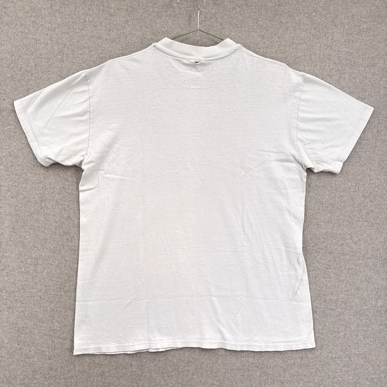 May include: A plain white short-sleeved t-shirt hanging on a metal hanger. The shirt is made of a soft, lightweight fabric and has a crew neck. The shirt is clean and appears to be in good condition.