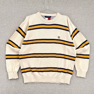 May include: Cream-colored sweater with horizontal stripes in navy blue and gold. The sweater has a crew neck and long sleeves. A small logo is visible on the chest. The sweater is hanging on a metal hanger.