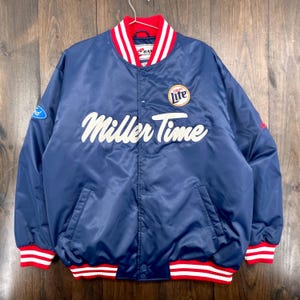 May include: A navy blue satin jacket with the words "Miller Time" in white script across the front. The jacket has a red and white striped collar, cuffs, and waistband. A "Miller Lite" logo patch is on the chest.