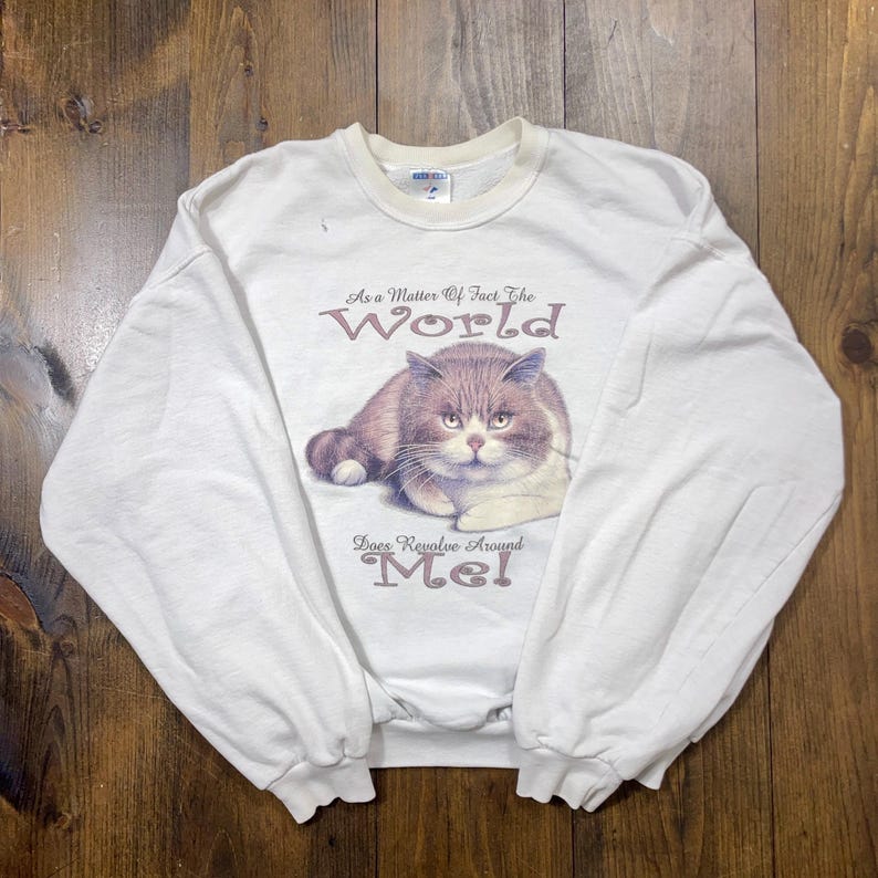 May include: White long-sleeve sweatshirt with a graphic of a brown and white cat. The text on the sweatshirt reads "As a matter of fact the world does revolve around me!"