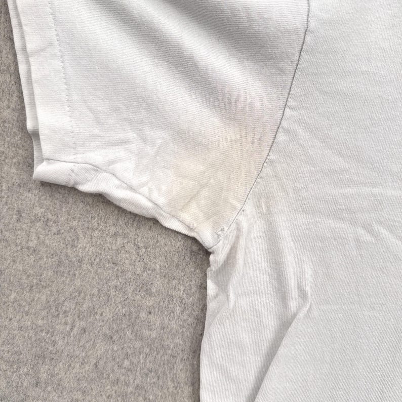 May include: Close-up of a light blue t-shirt sleeve, showing the shoulder and armpit area. The fabric is slightly wrinkled, and there are visible stains in the armpit area. The background is a light gray, textured surface.