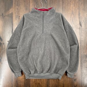May include: A gray fleece pullover with a half-zip closure and a red interior collar. The long-sleeved top is hanging on a metal hanger against a wooden background. The fleece material appears soft and warm.