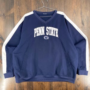 Puede incluir: Jersey azul marino con detalles blancos y el texto "PENN STATE" en letras blancas. El jersey tiene cuello en V y un pequeño bolsillo en la parte delantera. Las mangas tienen rayas blancas.