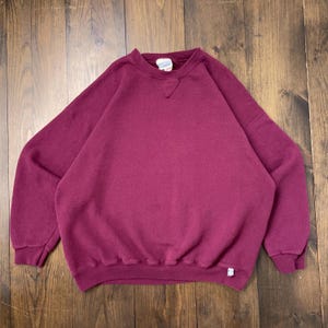 May include: A maroon crew neck sweatshirt with long sleeves. The garment has a relaxed fit and a small logo tag on the lower right. The sweatshirt is made of a soft, comfortable material, suitable for casual wear.