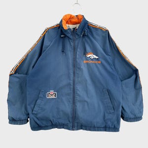 May include: A navy blue zip-up jacket featuring the Denver Broncos logo and text on the chest, an NFL Experience patch on the lower right pocket, and orange and white striped detailing running down the length of both sleeves with a high collar.
