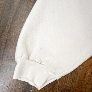 May include: Close-up of a cream-colored sweatshirt sleeve with a ribbed cuff. The fabric has a textured appearance, and there are a few small brown stains on the sleeve. The background is a dark wooden surface.