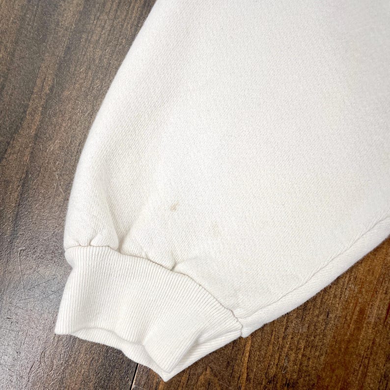 May include: Close-up of a cream-colored sweatshirt sleeve with a ribbed cuff. The fabric has a textured appearance, and there are a few small brown stains on the sleeve. The background is a dark wooden surface.